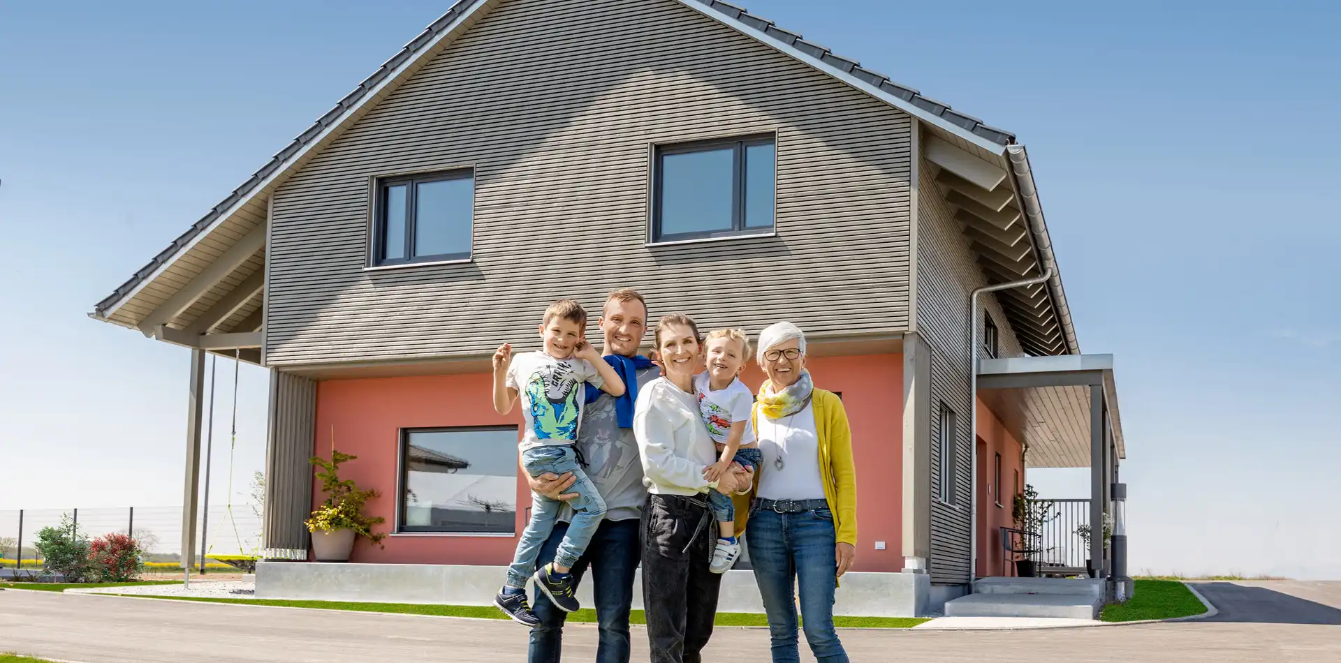 Familie aus drei Generationen vor einem modernen Haus mit Holzfassade im Obergeschoss und korallfarbener Fassade im Erdgeschoss.