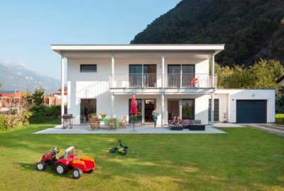 Ein modernes Einfamilienhaus mit großzügiger Terrasse auf der Vorderseite, eine Familie spaziert über gepflasterten Weg zum Hauseingang. Weiße und dunklere Fassadenelemente bilden einen klaren Kontrast, große Fensterflächen öffnen das Haus nach außen. Der Außenbereich ist gepflegt mit Rasen, Bepflanzung und einem kleinen Baum – vermittelt ein einladendes, modernes Modulhaus-Ambiente.